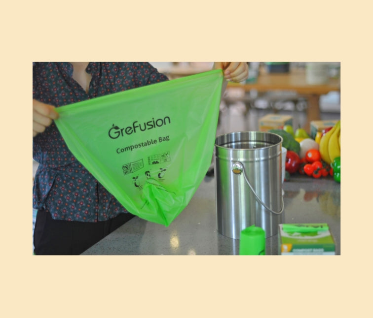 A person holds a green Grefusion compostable bag in a kitchen setting with a stainless steel container and various fruits and vegetables around.