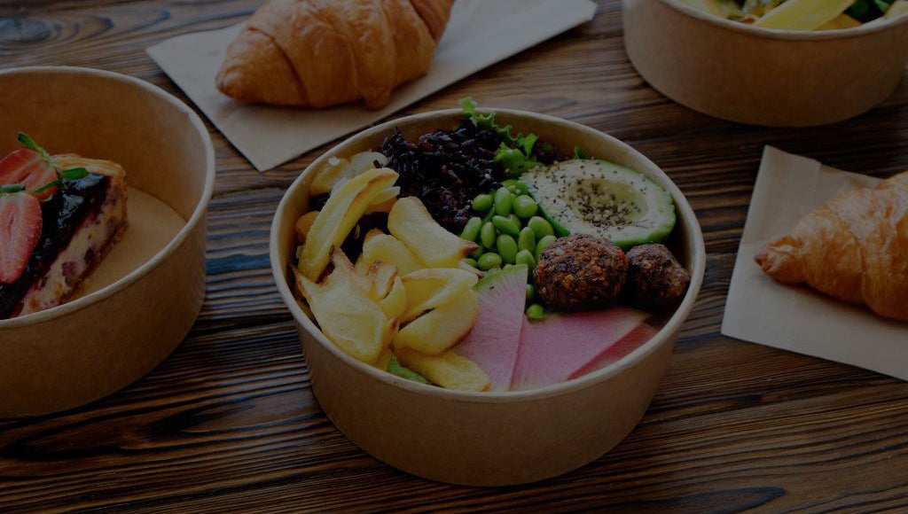 Colorful meals in paper bowls with avocado, edamame, rice, dessert, and croissant on wooden table; healthy food display.