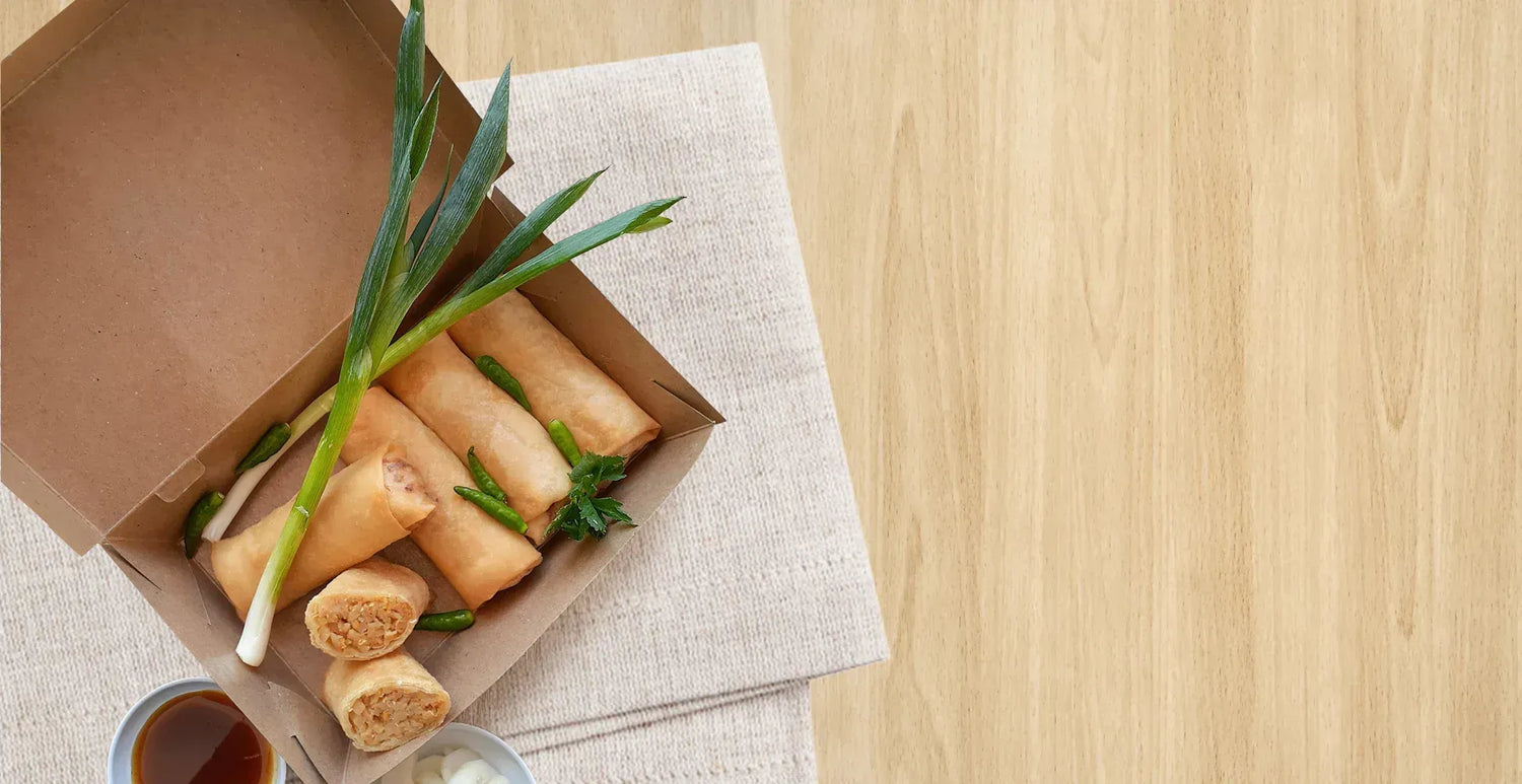 Golden spring rolls in a brown paper bowl, with dipping sauces, green onions, peppers, and cilantro.